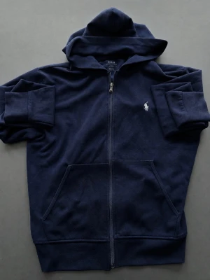 Mörkblå hoodie med dragkedja från Polo Ralph Lauren - Mörkblå hoodie från Polo Ralph Lauren med dragkedja framtill och klassisk vit broderad logga på bröstet. Väldigt bekväm. Står S fast passar med som en S-M.