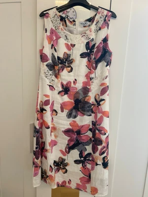 Blommig ärmlös klänning  - Snygg ärmlös klänning från Voglia i vitt med stora blommiga mönster i rosa, lila och svart. Klänningen har rund halsringning med spetsdetaljer och är gjord i ett luftigt linnematerial. Klänningen har en dragkedja på baksidan. Perfekt för varma dagar och ger en fräsch sommarkänsla.                                                  Är öppen för att förhandla priset🌸