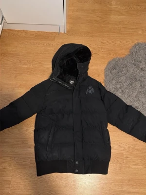 Svart pufferjacka från KWD - Säljer en svart pufferjacka från KWD med broderad logga på bröstet och huva. Jackan har ribbade muddar vid ärmslut och nederkant samt dragkedja och knappar framtill. Perfekt för kalla vinterdagar och riktigt skön att bära.
