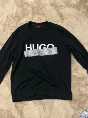 Svart sweatshirt från HUGO - Cool svart sweatshirt från HUGO med vit logga framtill och en unik silverfärgad tejpdetalj över bröstet. Tröjan har rund halsringning och långa ärmar. Perfekt för dig som gillar streetwear och vill sticka ut.