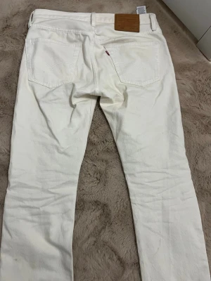 Vita raka jeans med knappgylf - Säljer ett par vita jeans med rak passform och knappgylf. Jeansen har klassiska fem fickor och bälteshällor. Materialet är kraftig denim och de har en enkel, clean look som passar till det mesta. Perfekta för dig som vill ha ett par stilrena och ljusa jeans.