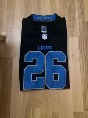 Svart NFL Detroit Lions tröja - Säljer en svart NFL Detroit Lions tröja från Nike med nummer 26 i blått och texten 'LIONS' på bröstet. Tröjan har blå detaljer på ärmarna och 'MOTOR CITY' i nacken. Perfekt för dig som gillar amerikansk fotboll och vill representera Lions.