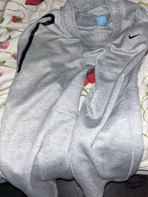 Gråa NOCTA Nike mjukisbyxor M - Gråa mjukisbyxor från Nike NOCTA med svart swoosh-logga och svart detalj längs ena benet. Byxorna har resår i midjan och är gjorda i mjukt material som är perfekt för chill dagar. Snygg och enkel design som passar till det mesta.