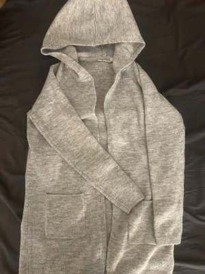 Grå stickad hoodiekofta - Mysig grå stickad kofta med huva och två framfickor. Långärmad modell i mjukt material, perfekt för lager på lager. Enkel och stilren design som passar till det mesta. Är i storlek 146/152 men skulle säga att den passar XS