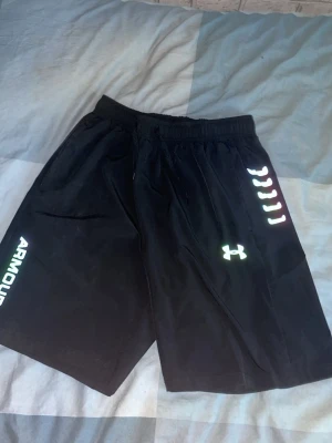 Svarta träningsshorts från Under Armour - Säljer ett par svarta träningsshorts från Under Armour med logga och reflekterande detaljer på sidan. Shortsen har elastisk midja och är tillverkade i ett lätt och snabbtorkande material, perfekta för sport och gym.