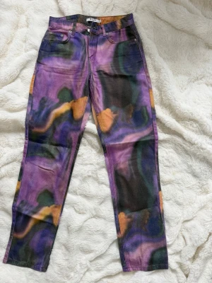 Färgglada raka jeans från NA-KD - Unika raka jeans från NA-KD med färgstarkt abstrakt mönster i lila, svart, orange och grönt. Klassisk femficksmodell med bälteshällor och knappgylf. Perfekta för dig som vill sticka ut med din stil.