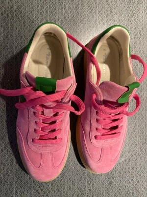 Rosa Tamaris sneakers med gröna detaljer - Snygga sneakers från Tamaris i rosa mocka med gröna detaljer och rosa skosnören. Skorna har en beige sula och en klassisk låg siluett. Perfekta för dig som vill sticka ut med färg och stil. Märket syns på sidan i vitt. Skorna bara använda en dag, köpte fel storlek! Nypris 1200kr