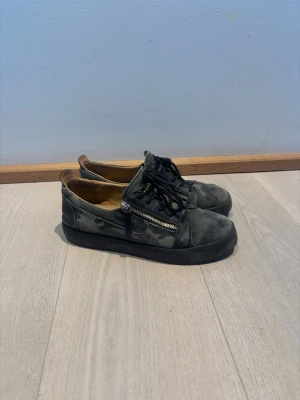 Camo Giuseppe Zanotti skor - Snygga Zanottis i grönt camomönster med svarta snören och guldig dragkedja på sidan. Skorna har låg profil och svart sula. Materialet ser ut att vara mocka eller liknande textil. Perfekta för en Laidback Grisch stil