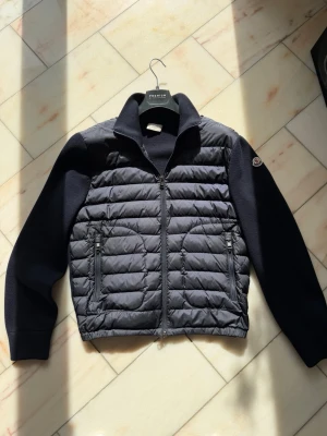 Moncler cardigan  - Snygg mörkblå cardiganjacka med quiltad front och stickade ärmar. Jackan har dragkedja framtill, två fickor med dragkedja och en hög krage. Moncler-logga på ena ärmen. Perfekt för lager-på-lager och stilren look.