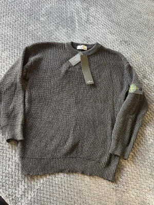 Mörkgrå stickad tröja Stone Island - Säljer en mörkgrå stickad tröja från Stone Island med klassisk patch på ärmen. Tröjan har rund hals, ribbade muddar och är gjord i ett grovt stickat material. Perfekt för dig som gillar streetwear och vill ha något stilrent och bekvämt.