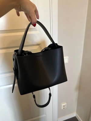 Svart Arket handväska  - Säljer denna snygga populära handväskan från Arket. Den har både ett långt axelremsband men också ”handtaget”. Den är som ny och endast använd några fåtal gången