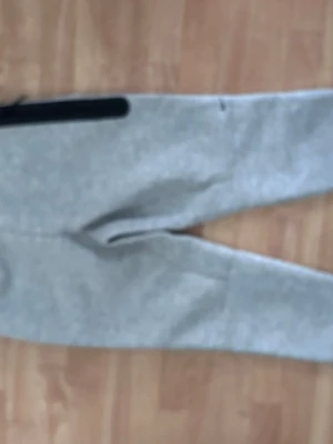 Gråa mjukisbyxor från Nike - Snygga gråa mjukisbyxor från Nike med svart logga på benet. Byxorna har dragsko i midjan och praktiska fickor med dragkedja. Tillverkade i mjukt och bekvämt material som passar perfekt för chill eller träning.