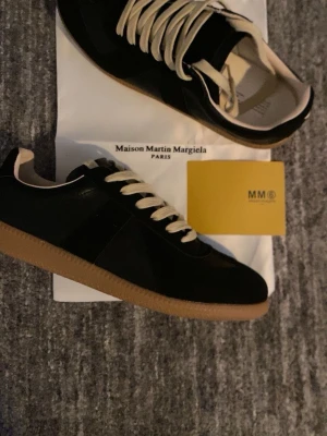 Maison Margiela svarta sneakers - Svarta sneakers från Maison Martin Margiela med beige detaljer och klassiska vita snören. Skorna är i riktigt bra kvalité, de är helt nya. Skorna är för dig som är ute efter en snygg stil och vill ha något att extra.