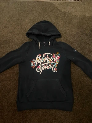 Mörkblå hoodie Superdry med blommigt tryck - Svart hoodie från Superdry med stor blommig logga på bröstet och vita dragsnören i huvan. Klassisk känguruficka framtill och ribbade muddar. Perfekt för en avslappnad och trendig stil.