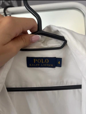 Vit skjorta från Polo Ralph Lauren - Klassisk vit skjorta från Polo Ralph Lauren med blå broderad logga på bröstet. Skjortan har knappar framtill och är tillverkad i mjuk bomull. Perfekt till både jeans och kostymbyxor för en clean och stilren look.