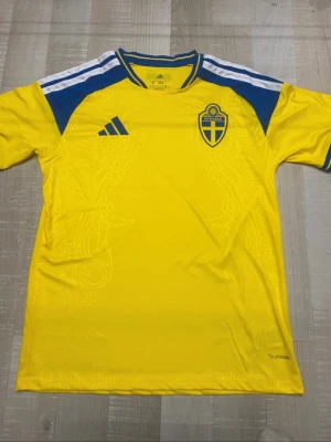 Svensk landslagströja Adidas gul/blå - Gul svensk landslagströja från Adidas med blå detaljer och vita ränder på axlarna. Broderat landslagsemblem på bröstet och 'Sverige' tryckt i nacken. Tillverkad i lätt och ventilerande funktionsmaterial, perfekt för fotboll eller träning.