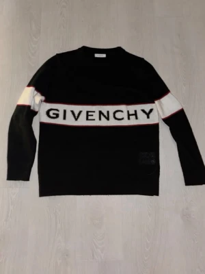 Svart stickad tröja från Givenchy - Snygg svart stickad tröja från Givenchy med vit panel och stor logga över bröstet. Tröjan har långa ärmar, rund halsringning och röda detaljer längs panelen. Perfekt för dig som gillar streetstyle och exklusiva märken. 
