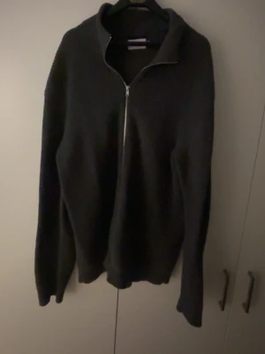 Grå stickad tröja med dragkedja - Mysig svart stickad tröja med hög krage och dragkedja framtill. Tröjan har lång ärm och är tillverkad i ett tjockare stickat material som passar perfekt när det är kallt. Enkel och stilren design utan synliga loggor eller mönster.