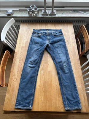 Lean Dean Nudie jeans - Säljer ett par klassiska blå jeans med raka ben och lätt slitna detaljer vid knän och bakfickor. Jeansen har fem fickor, normal midja och är tillverkade i denim. Perfekta för en avslappnad och trendig look.