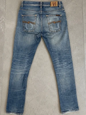 Nudie Jeans 30/32 - Säljer ett par ljusblå jeans med snygga slitningar och tvättade detaljer. Jeansen har klassiska fem fickor, kontrastsömmar och en patch bak i midjan. Materialet är denim i bomull.