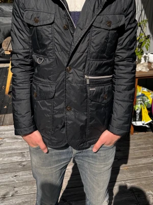 Svart quiltad jacka från Snoot - Svart quiltad jacka från Snoot med flera fickor framtill, både med knapp och dragkedja. Jackan har en klassisk krage, knappstängning och lång ärm. Materialet är glansigt och lätt, perfekt för kyligare dagar. Tveka inte att höra av dig vid minsta fråga eller fundering 🙌🙏