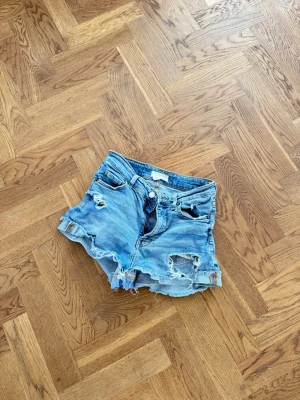 Blå slitna jeansshorts med fransar - Säljer ett par blå jeansshorts med slitna detaljer och fransig kant. Shortsen har klassisk femficksdesign, bälteshällor och knappgylf. Perfekta för en avslappnad och trendig look.