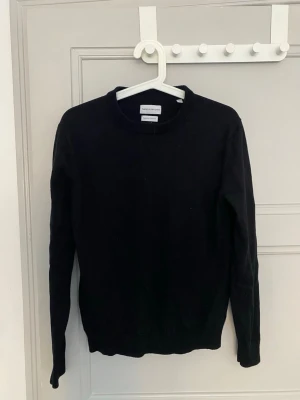 Svart stickad tröja med rund hals - En enkel och stilren svart stickad tröja med rund hals och långa ärmar. Tröjan har en klassisk passform och ribbade muddar vid ärmslut och nederkant. Perfekt basplagg till garderoben.
