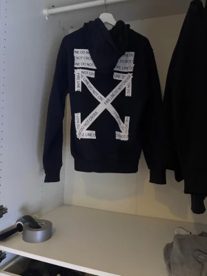 Marinblå hoodie med tryck - Säljer en svart hoodie med stor huva och känguruficka. På ryggen finns ett stort vitt tryck med texten 'DO NOT CROSS' i ett korsmönster och framtill står det 'OFF' samt en detalj med samma text. Perfekt för dig som gillar streetwear och tydliga prints.