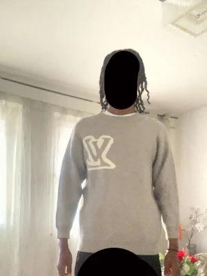 LV stickad tröja  - Säljer en stilren grå tröja med stor vit LV-logga på bröstet. Tröjan har rund halsringning och långa ärmar. Perfekt för dig som gillar enkel men snygg design.