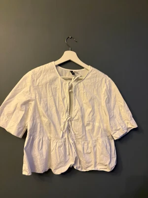 Vit blus med knytning från H&M Divided - Söt vit blus från H&M Divided med korta ärmar och knytning framtill. Perfekt att matcha med jeans eller kjol för en fräsch stil.
