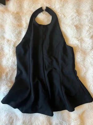 Svart halterneck topp - Snygg svart halterneck topp med knäppning i nacken och utsvängd passform. Tillverkad i ett mjukt, slätt tyg som ger ett elegant intryck. Perfekt för dig som vill ha en stilren och trendig look.