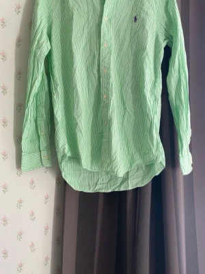 Grön- och vitrandig skjorta Polo Ralph Lauren - Snygg långärmad skjorta från Polo Ralph Lauren i grönt och vitt randigt bomullstyg. Klassisk krage, knäppning framtill och lila broderad logga på bröstet. Perfekt för en fräsch och stilren look.