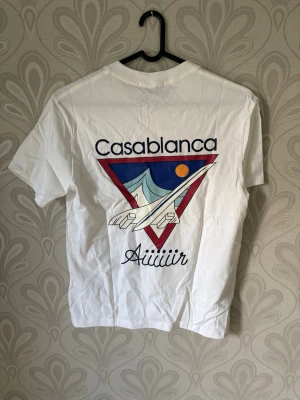 Vit Casablanca t-shirt med tryck - Vit t-shirt från Casablanca med stort grafiskt tryck på ryggen som visar ett flygplan, berg och sol i färgerna blå, röd och orange. Framtill står det 'Aiiiiiir' och Casablanca-loggan syns både på bröstet och etiketten. Klassisk passform och rund hals.