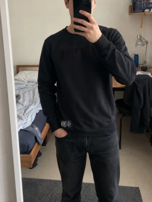 Svart sweatshirt med New York-tryck - Säljer en svart sweatshirt med texten 'New York' på bröstet och en liten patch vid nederkanten. Tröjan har rund hals, långa ärmar och ribbade muddar. Perfekt för dig som gillar enkel och stilren streetwear.