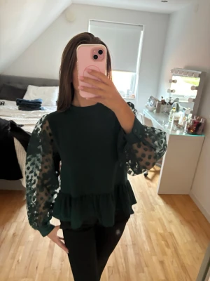Mörkgrön topp med volang och puffärm - Trendig mörkgrön topp med transparenta puffärmar i mesh och prickigt mönster. Toppen har volang nedtill och en loose passform som ger en cool och avslappnad look. Perfekt för dig som vill sticka ut lite extra.