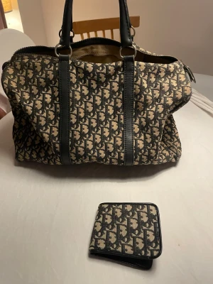 Dior vintage ! En av dom första  - Säljer en stor resväska och matchande plånbok från Dior med klassiskt monogrammönster i beige och svart. Väskan har svarta skinnhandtag och detaljer, dragkedja och rymligt innerfack. Plånboken har samma mönster och svarta kanter. Perfekt för dig som gillar lyxiga accessoarer.     D en luktar gammal mormor men den e från vår gammelmormors tid. Kan mötas i pantbank osv 