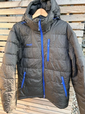 Svart dunjacka med blå dragkedjor - Säljer en svart dunjacka från Bergans med blå detaljer och dragkedjor. Jackan har huva, tre framfickor med dragkedja och quiltad design. Perfekt för kalla vinterdagar och riktigt skön att bära. 