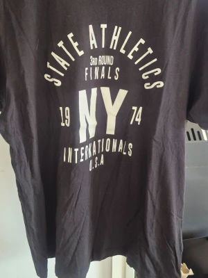 Svart t-shirt med vit text NY - Säljer en svart t-shirt med vit tryckt text och grafik på framsidan där det står 'STATE ATHLETICS', 'NY', 'INTERNATIONALS U.S.A.' och siffrorna 1974. T-shirten har rund hals och korta ärmar. Perfekt för en sportig och avslappnad stil.