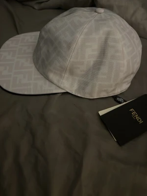 Vit keps med Fendi-logga - Säljer en stilren vit keps från Fendi med det klassiska FF-monogrammet över hela kepsen. Kepsen har böjd skärm och är tillverkad i ett tygmaterial som känns exklusivt. Perfekt accessoar för dig som gillar designerplagg och vill sticka ut.