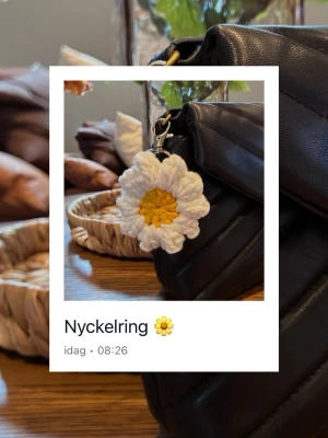 Virkad nyckelring  - Söt handgjord nyckelring i form av en blomma, virkad i vitt och gult garn. Perfekt att hänga på väskan eller använda till nycklar. Blomman har en mjuk och fluffig känsla och fästs enkelt med en metallknippet.