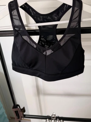 Svart sport-bh med meshdetaljer - Snygg svart sport-bh med bred resår och meshdetaljer både fram och bak. Bh:n har brottarrygg och hakknäppning bak för extra stöd. Tillverkad i stretchigt material som andas, perfekt för träning eller en aktiv vardag.