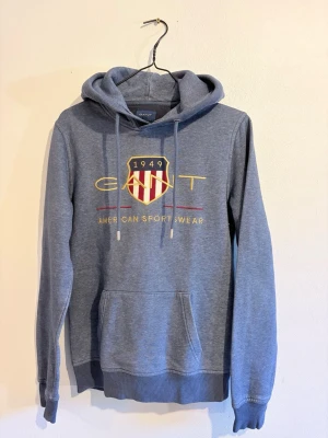Blå hoodie från GANT med tryck - Snygg blå hoodie från GANT med klassiskt tryck och logga framtill. Hoodien har snörning i huvan, känguruficka och ribbade muddar. Tillverkad i mjuk bomullsmix som är perfekt för chill dagar. Passar dig som gillar sportig och clean stil.