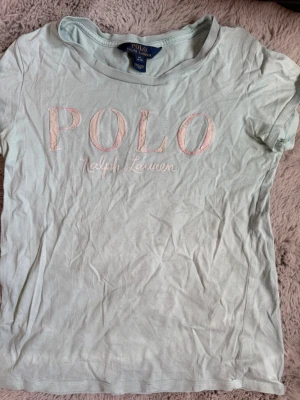 Turkos Polo Ralph Lauren t-shirt - Snygg turkos t-shirt från Polo Ralph Lauren med broderad logga i rosa och vitt framtill. Klassisk rund halsringning och korta ärmar. Perfekt för en avslappnad och stilren look. Storlek m i barnstorlek. 