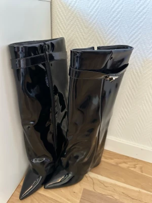 Svarta boots - Säljer ett par svarta boots i glansigt lackmaterial med spetsig tå och dekorativ rem upptill i storlek 37 men passar mig som  vanligtvis har storlek 38. Dragkedja på insidan för enkel påtagning. Snygg och trendig modell.💕
