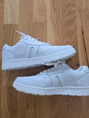 Vita sneakers från J.Lindeberg - Stilrena vita sneakers från J.Lindeberg med klassisk låg profil. Skorna har vita snören, perforerad tå för ventilation och diskreta sömdetaljer. Ovandelen är i slätt syntetmaterial och sulan är vit med bra grepp. Perfekta för en clean och modern look.