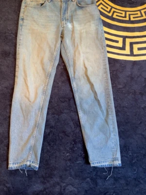 Ljusblå raka jeans med hög midja - Säljer ett par ljusblå jeans med rak passform och hög midja. Jeansen har klassisk femficksdesign, gylf med dragkedja och knapp samt råa kanter vid bensluten för en avslappnad look. Perfekta för dig som gillar vintagekänsla och enkel stil.