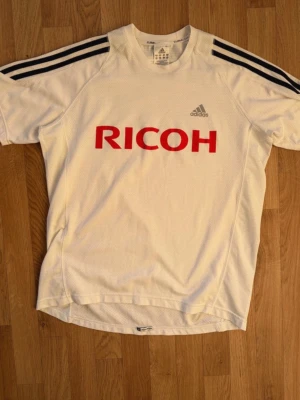 Vit Adidas Träningströja - Säljer en vit träningströja från Adidas med Climalite-material. Tröjan har svarta ränder på axlarna, stor röd RICOH-logga på bröstet och Adidas-logga i grått. Perfekt för träning eller sportaktiviteter.