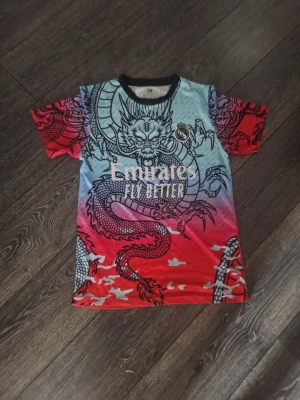 Färgstark Real Madrid t-shirt drake - Säljer en unik och färgstark Real Madrid t-shirt med stort drakmotiv och texten 'Emirates Fly Better' på bröstet. T-shirten har korta ärmar och en cool mix av rött, blått och svart. Perfekt för dig som vill sticka ut med en sportig och kreativ look.
