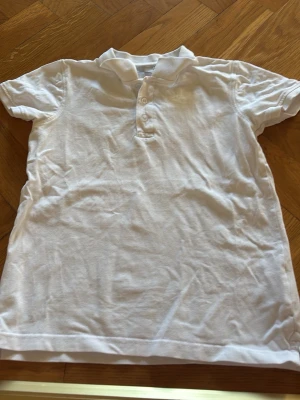 Vit kortärmad T-shirt från Lager 157 i stl 140 - Klassisk vit polotopp med korta ärmar och knappar framtill. Perfekt till jeans  för en clean och enkel stil. Tillverkad i mjukt bomullsmaterial som känns skönt mot huden. Den är ifrån Lager 157 i stl 140. Jättebra skick. 
