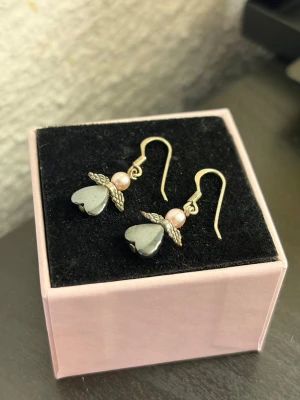 Hjärthängande örhängen med pärlor - Söta örhängen med metallkrokar, dekorerade med ljusrosa pärlor och hjärtformade hängen i mörkgrå metall som ska föreställa en ängel. 